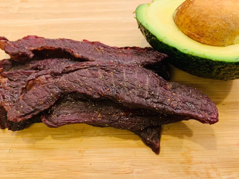 Long strips of homemade beef jerky beside an avocado half