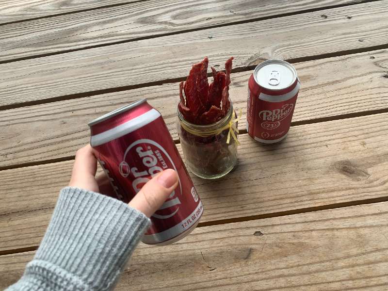 A hand holding a Dr Pepper can beside jerky and another can