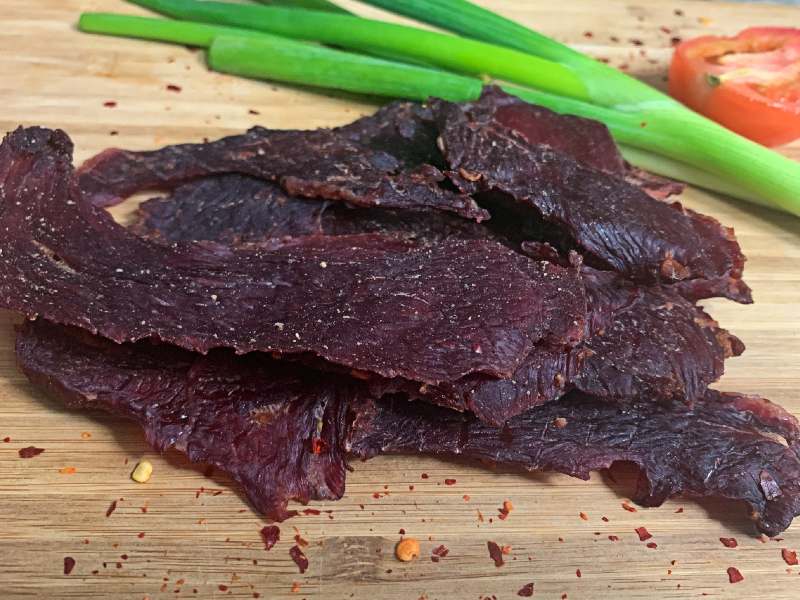 Wide spicy beef jerky pieces beside scallions and a tomato