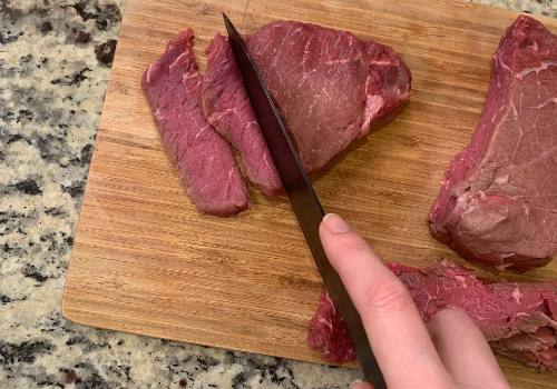 A knife cutting beef into thin slices on a board