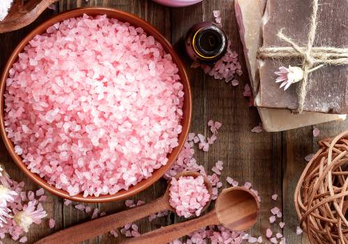 Coarse pink crystals in a wooden bowl beside small bottles