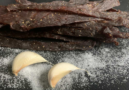 Homemade beef jerky beside garlic cloves on a dark surface