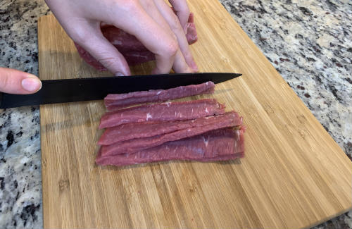 Thin raw beef strips being cut on a wooden board
