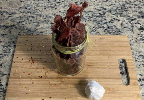 A glass jar of finished jerky on a cutting board beside garlic