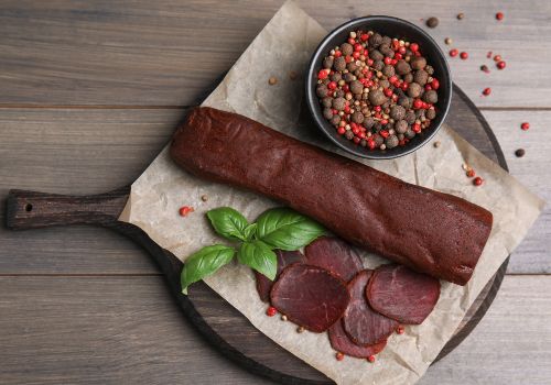 A dried meat roll with slices beside peppercorns and herbs