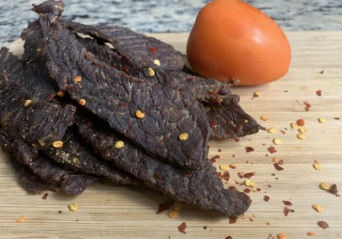 Pepper-flecked beef jerky on a board beside a tomato