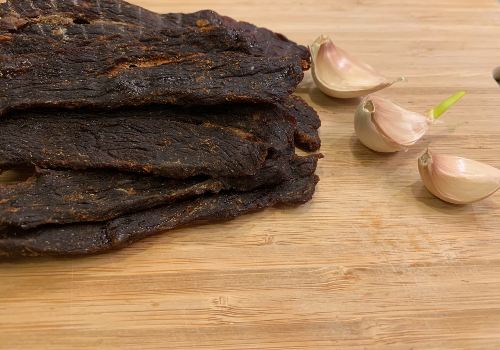 Thick beef jerky strips on a wooden board beside garlic cloves